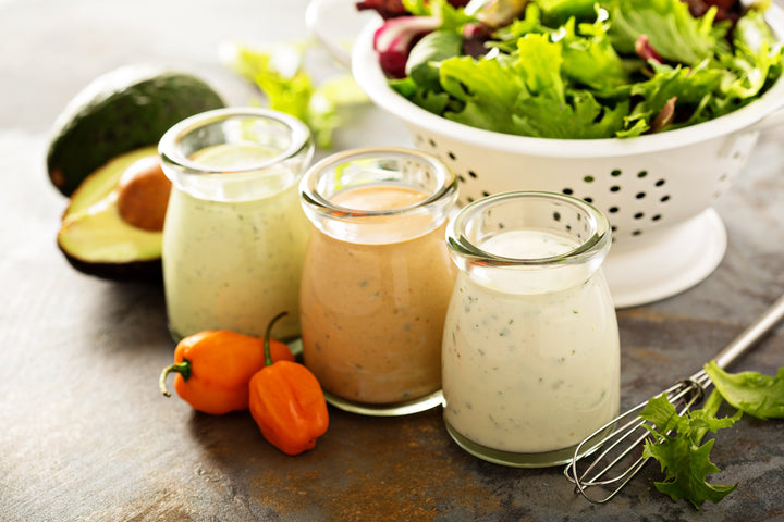 Drei Dressings in Gläsern neben einer Schüssel Salat, Avocado und Paprika